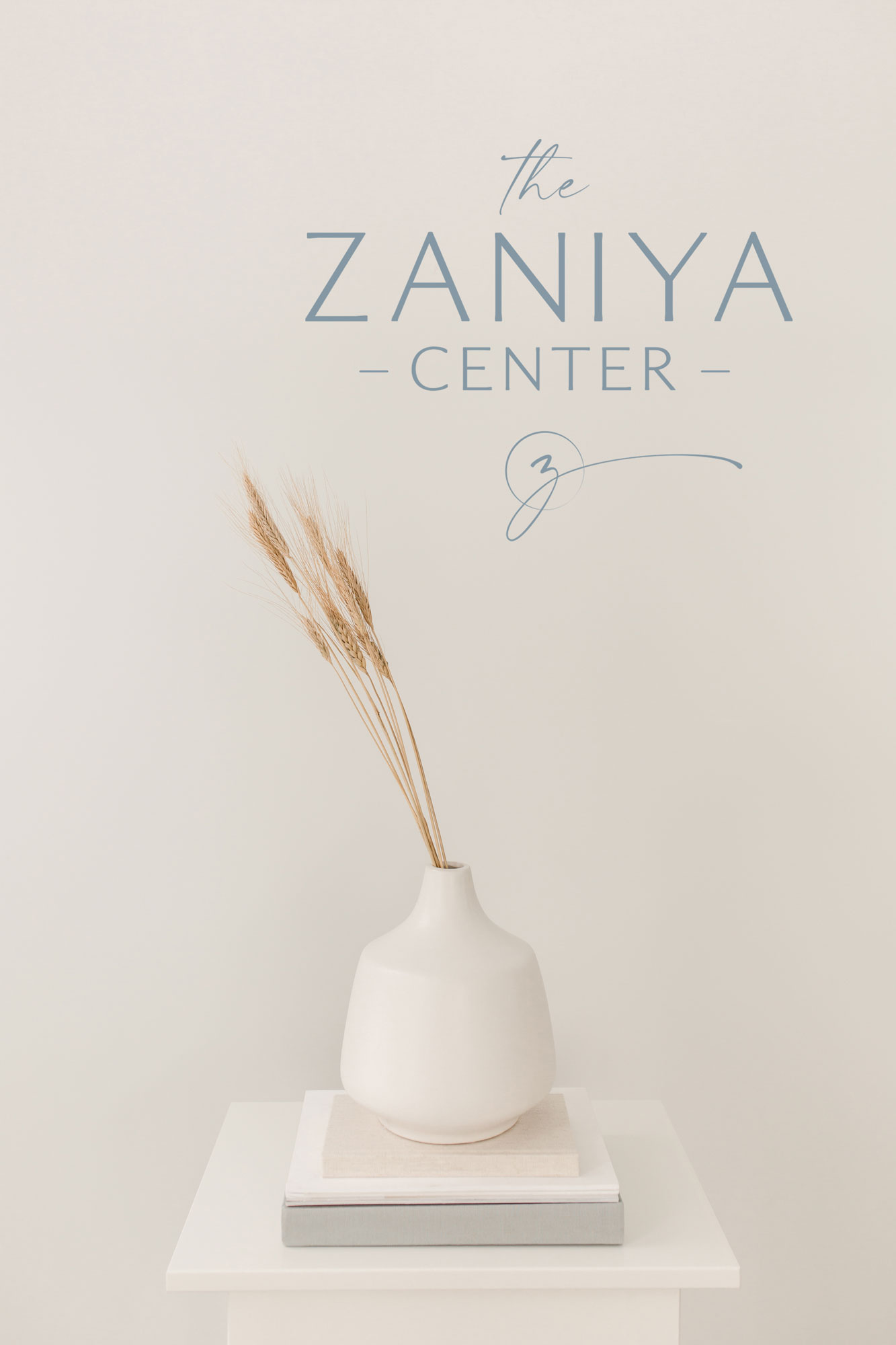 Stack of books on a white end table with a ceramic vase of tall grasses, the wall in the background features the Zaniya Center logo.
