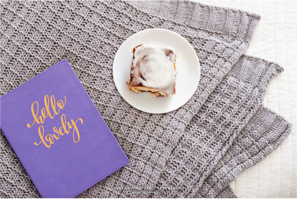 Iced cinnamon roll on a plate sitting on a knit blanket next to a book titled "Hello Lovely".

