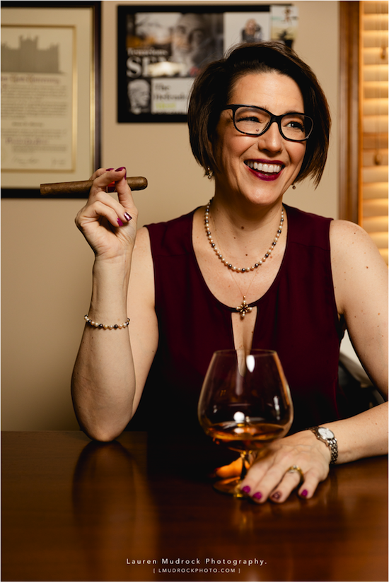 Woman wearing bright red lipstick 
sitting at a table with a glass of brandy in one hand and a cigar in the other.
