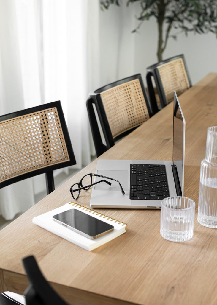Long wooden table with laptop, planner, phone, eye glasses and glass of water.
